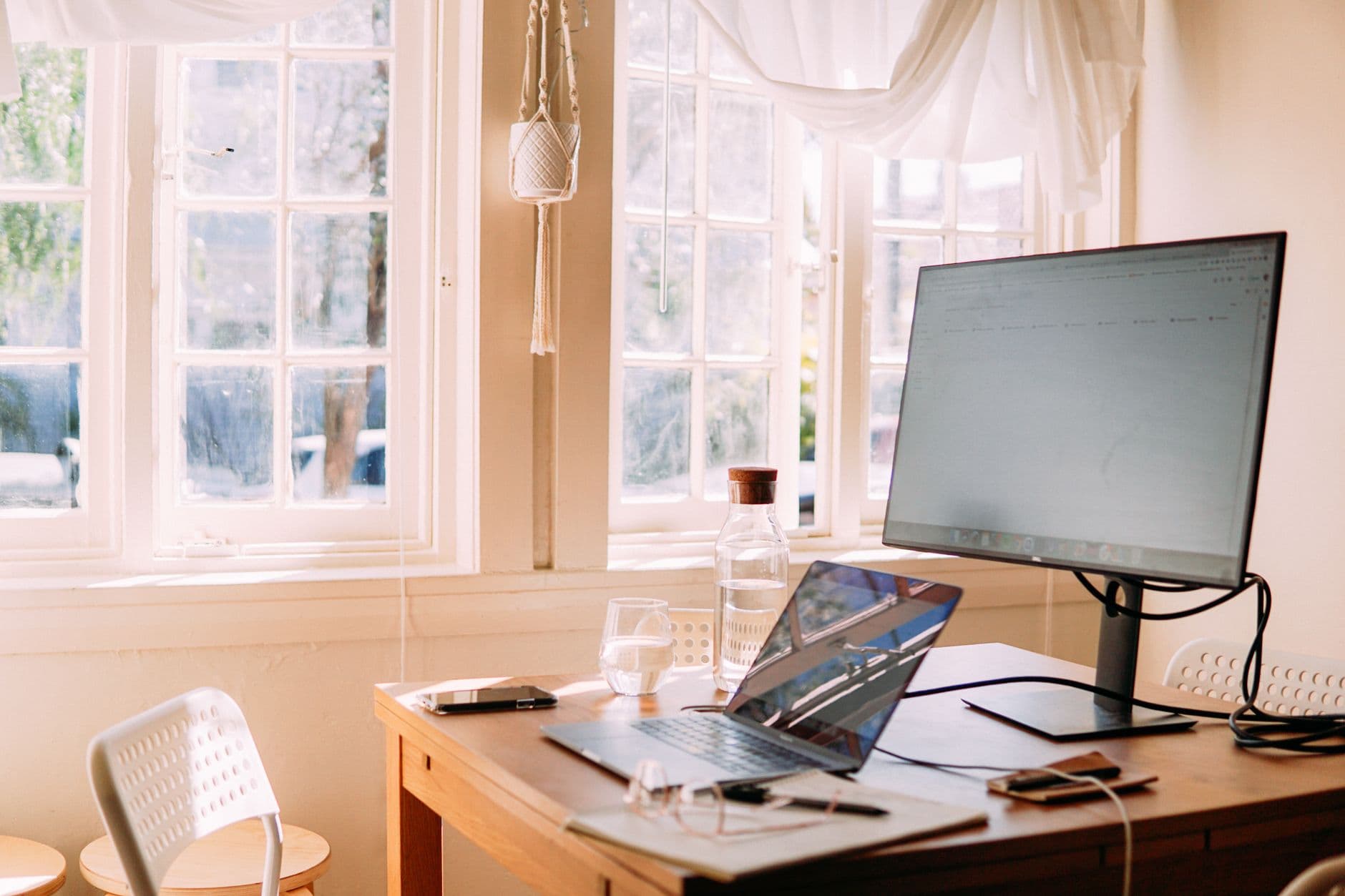 A workspace with a laptop on a wooden desk, daylight through a large window.